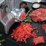 Carne cruda de res dispuesta sobre una mesaa al lado de una moledora de carne
